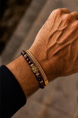Gold mesh bracelet stacked in front of a red Tiger Eye bead bracelet with a gold Buddha accent on a masculine wrist.