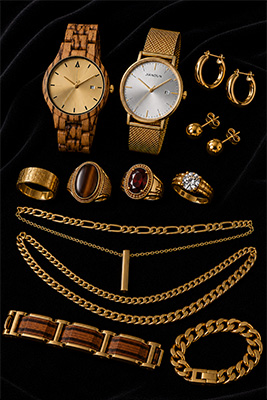 Warm coordinated men's jewelry collection on black velvet using gold, zebrawood, Tiger Eye, marble beads, a wooden watch, bracelets, chain, ring, and earrings.